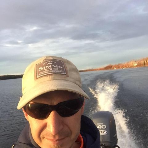 man in sunglasses and a hat taking a selfie on a boat with water and tree lined shore in the background