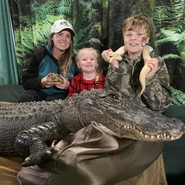 woman and two children posing with a large alligator, a turtle, and a snake in a jungle themed setting